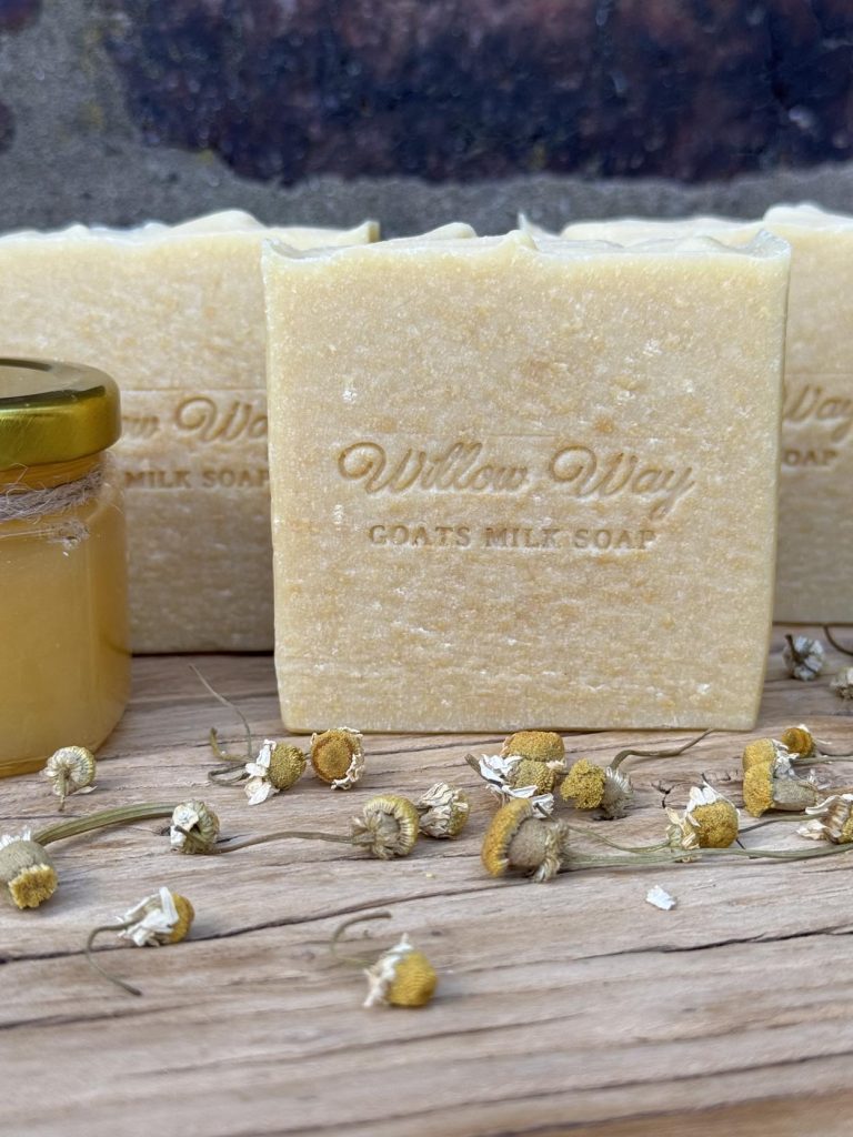 A bar of goats milk soap on a wooden board with honey and roman chamomile