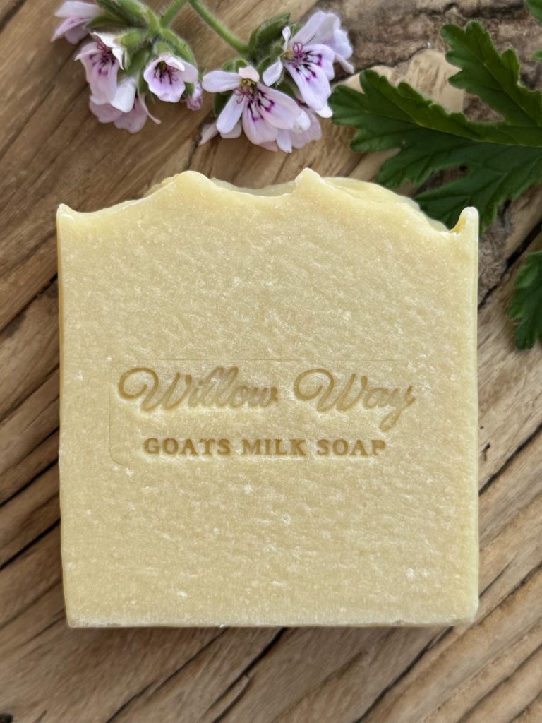 A bar of goats milk soap on a wooden board with rose geranium and clary sage